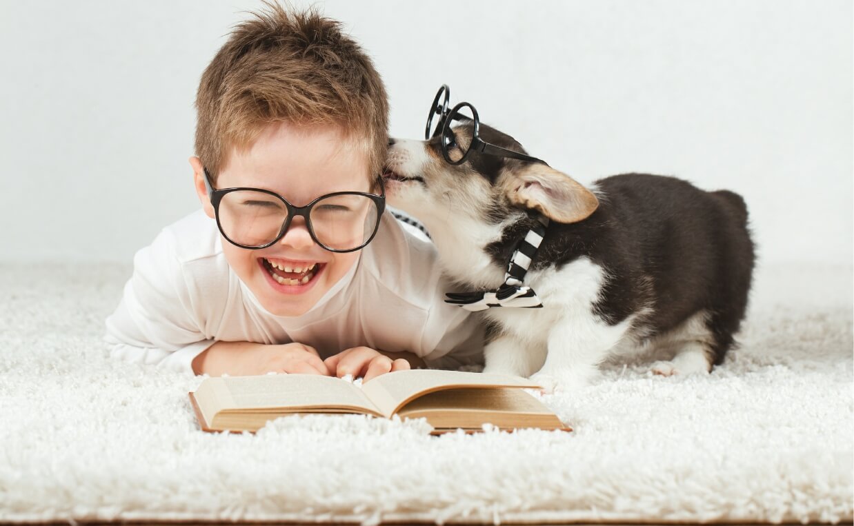 cute young boy with big black reading glasses and book in front of him laughing while getting licked by his puppy
