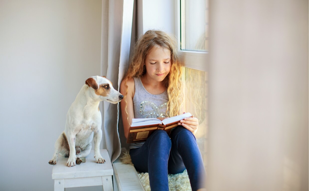 elementary age girl sitting by window reading to jack russell terrier dog